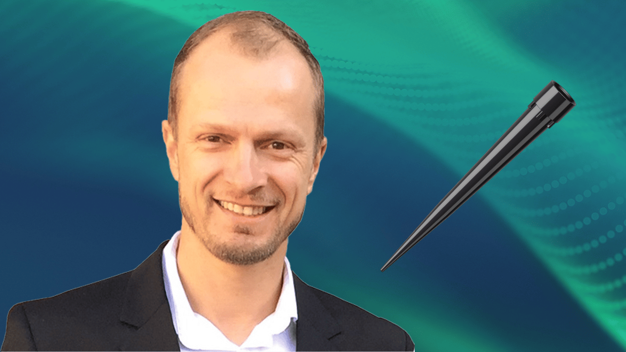 Smiling person in a dark blazer beside a black laboratory pipette tip, on a teal gradient background.