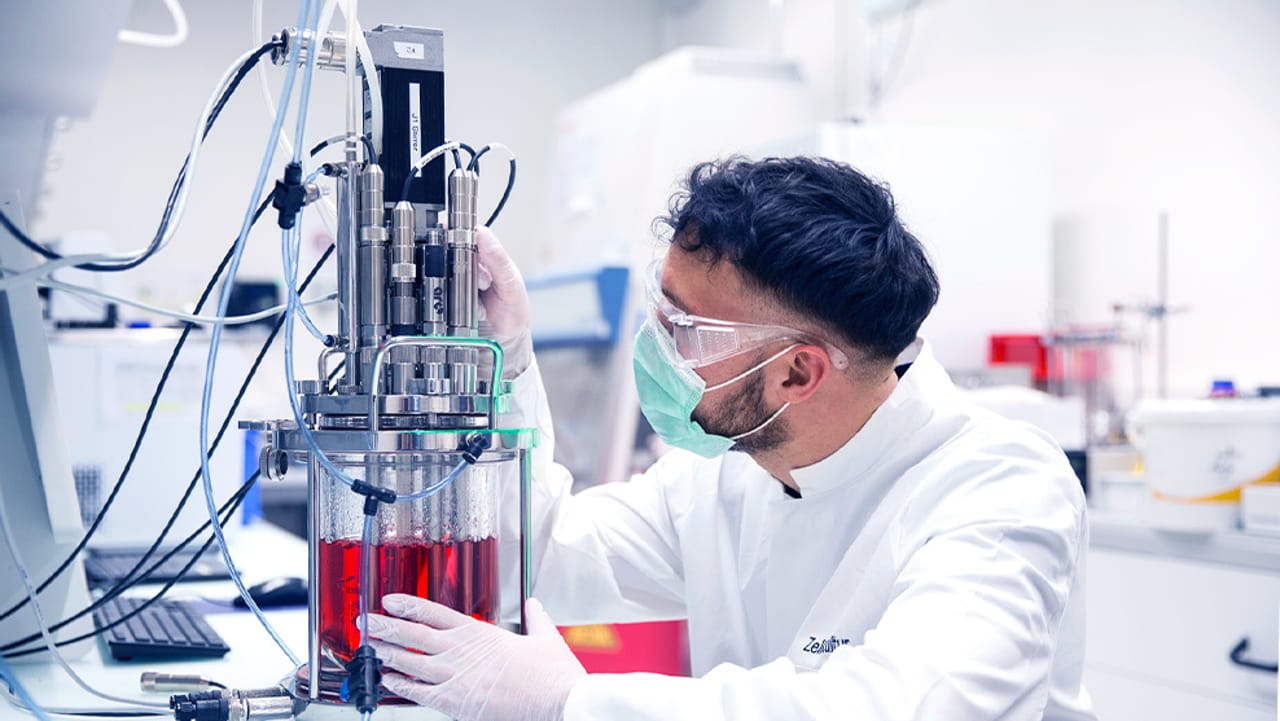 Lab technician in white coat, safety goggles, and mask adjusting a bioreactor with red cell culture media.