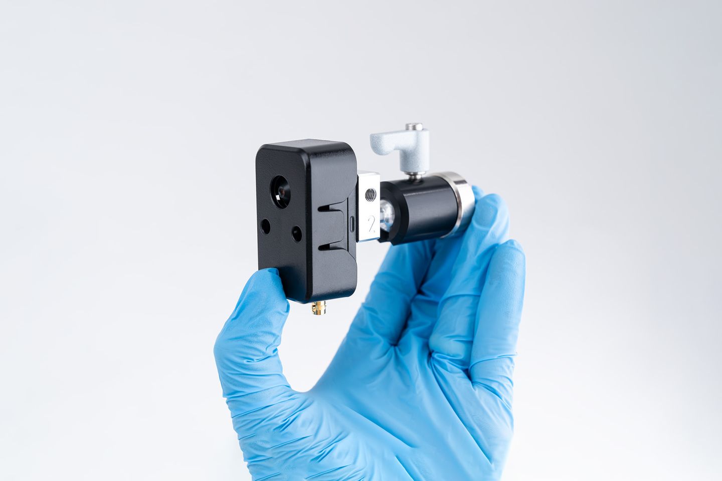 Gloved hand holding a small black rectangular camera module with lens ports attached to a cylindrical mount with adjustment knob