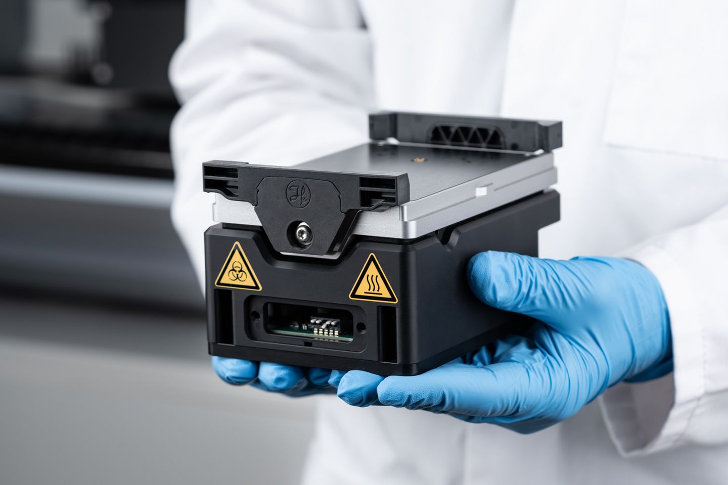 Gloved hands holding a black modular liquid handling head with biohazard and hot surface warning labels
