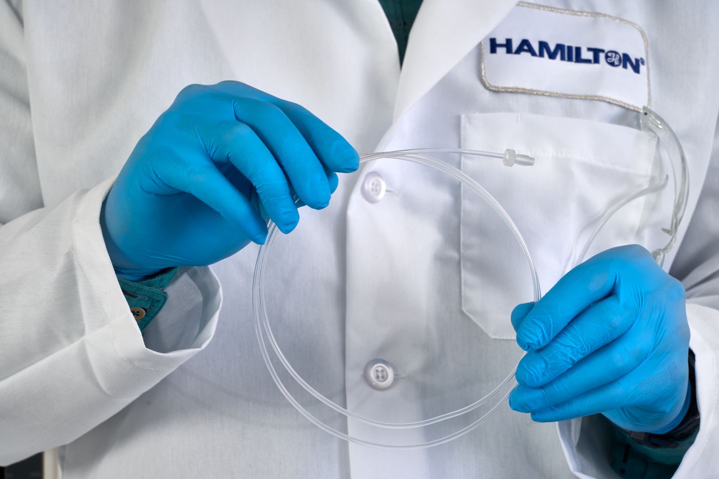 Lab coat-wearing person in blue nitrile gloves holding a clear flexible tubing loop with a luer fitting