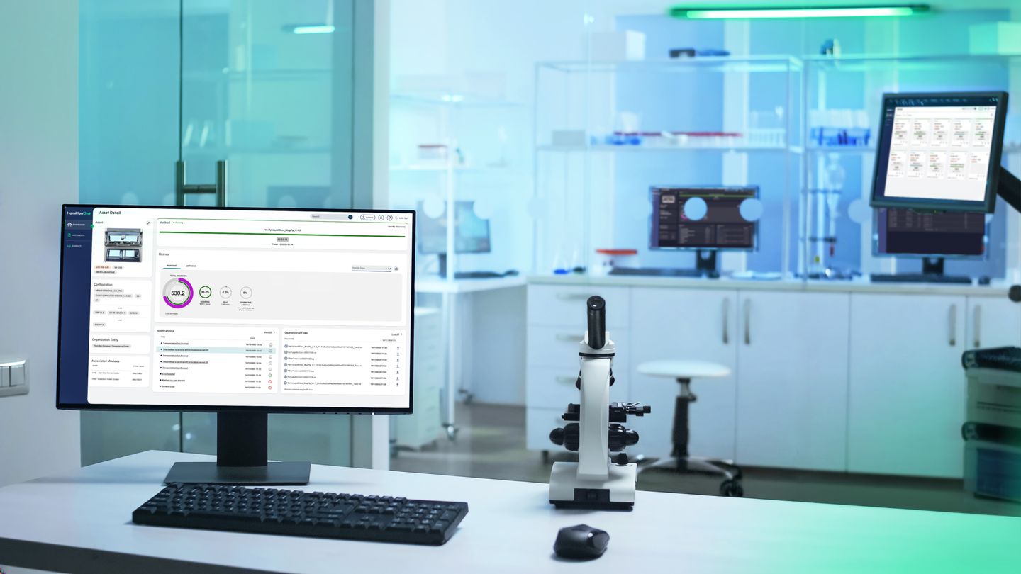 Laboratory workstation with a desktop monitor displaying a Hamilton asset management dashboard, alongside a microscope and keyboard.