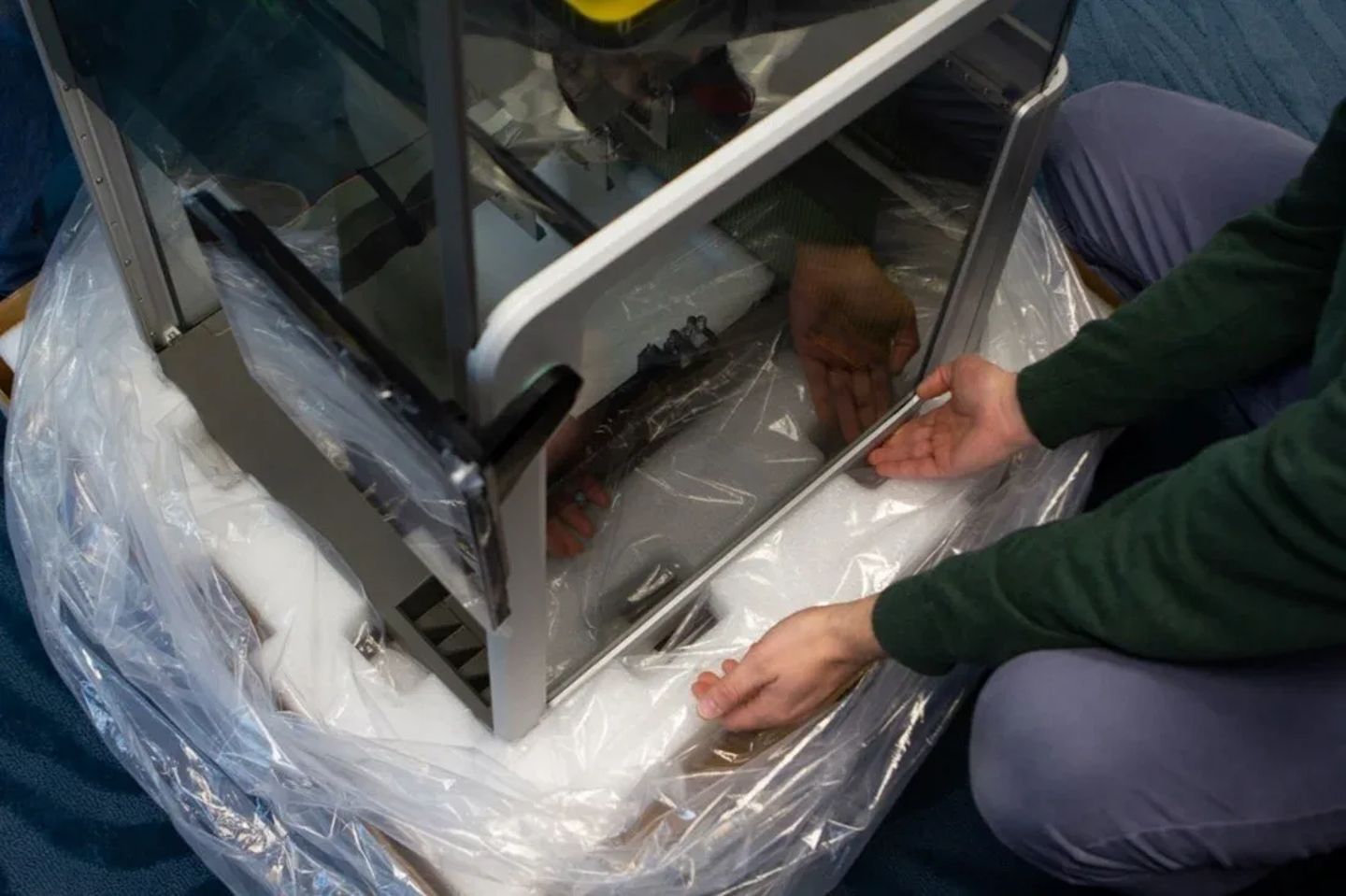 Person unboxing a laboratory instrument with metal frame and clear panels from protective packaging
