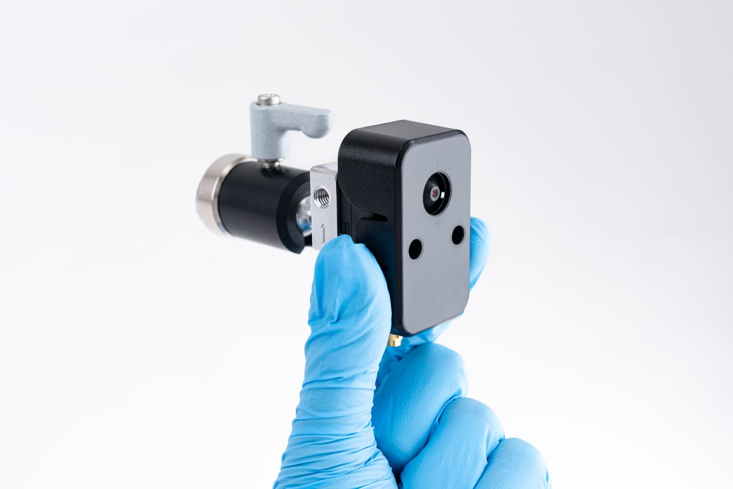 Gloved hands holding a compact camera module with lens assembly against a white background