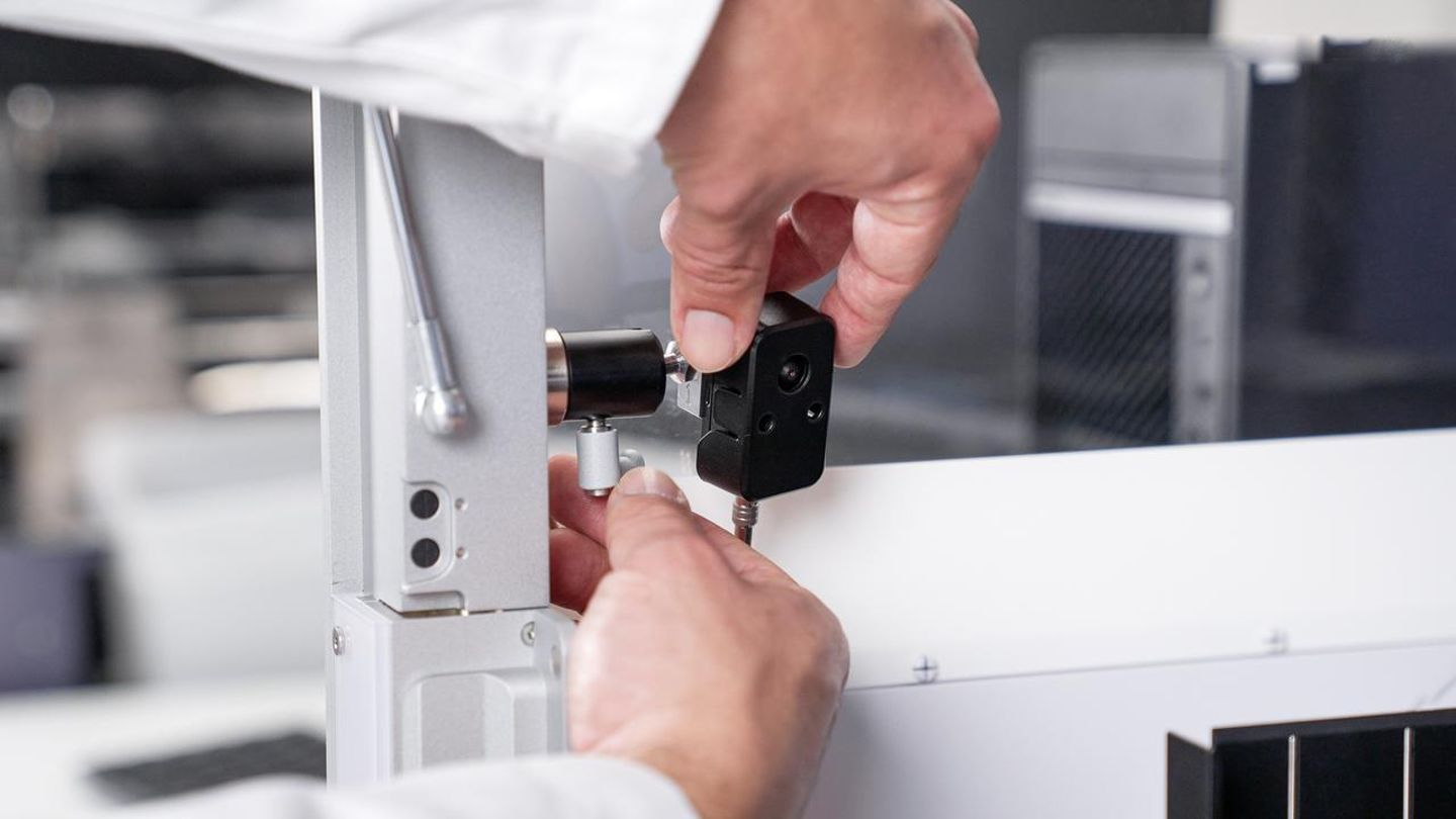 Hands mounting a small black camera module onto a white laboratory instrument frame