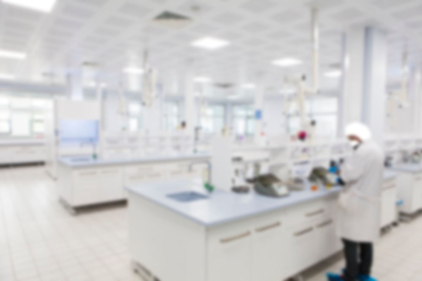 Laboratory with white benches and equipment; one person in lab coat and cap working at a central bench.
