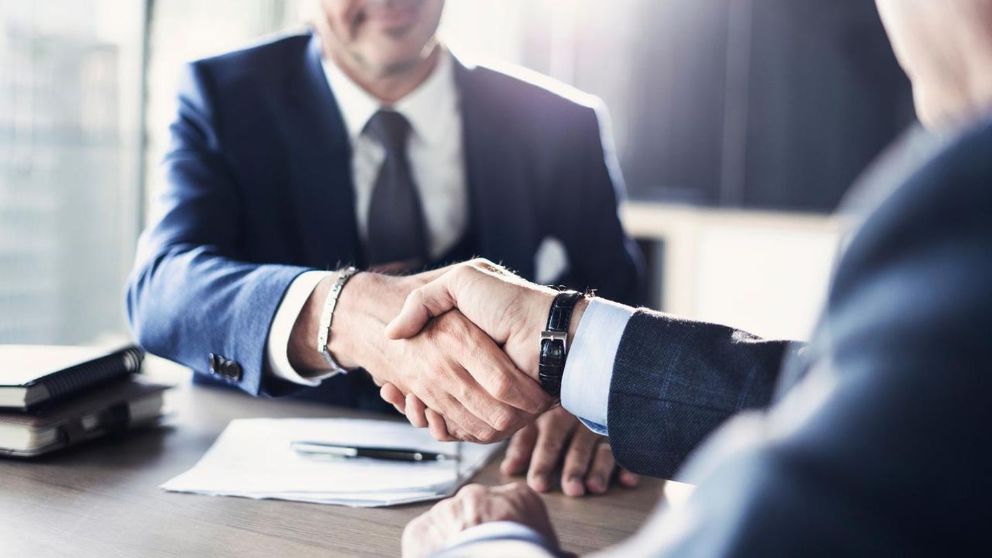 Two professionals in suits shaking hands across a desk with documents and folders nearby.