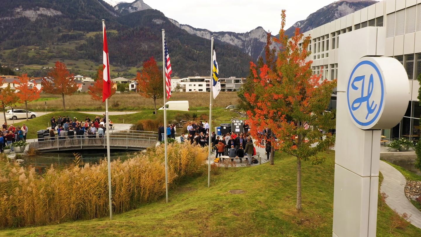 Outdoor gathering at Hamilton Bonaduz AG campus with Alpine mountains, autumn trees, and three flags including Swiss and US.