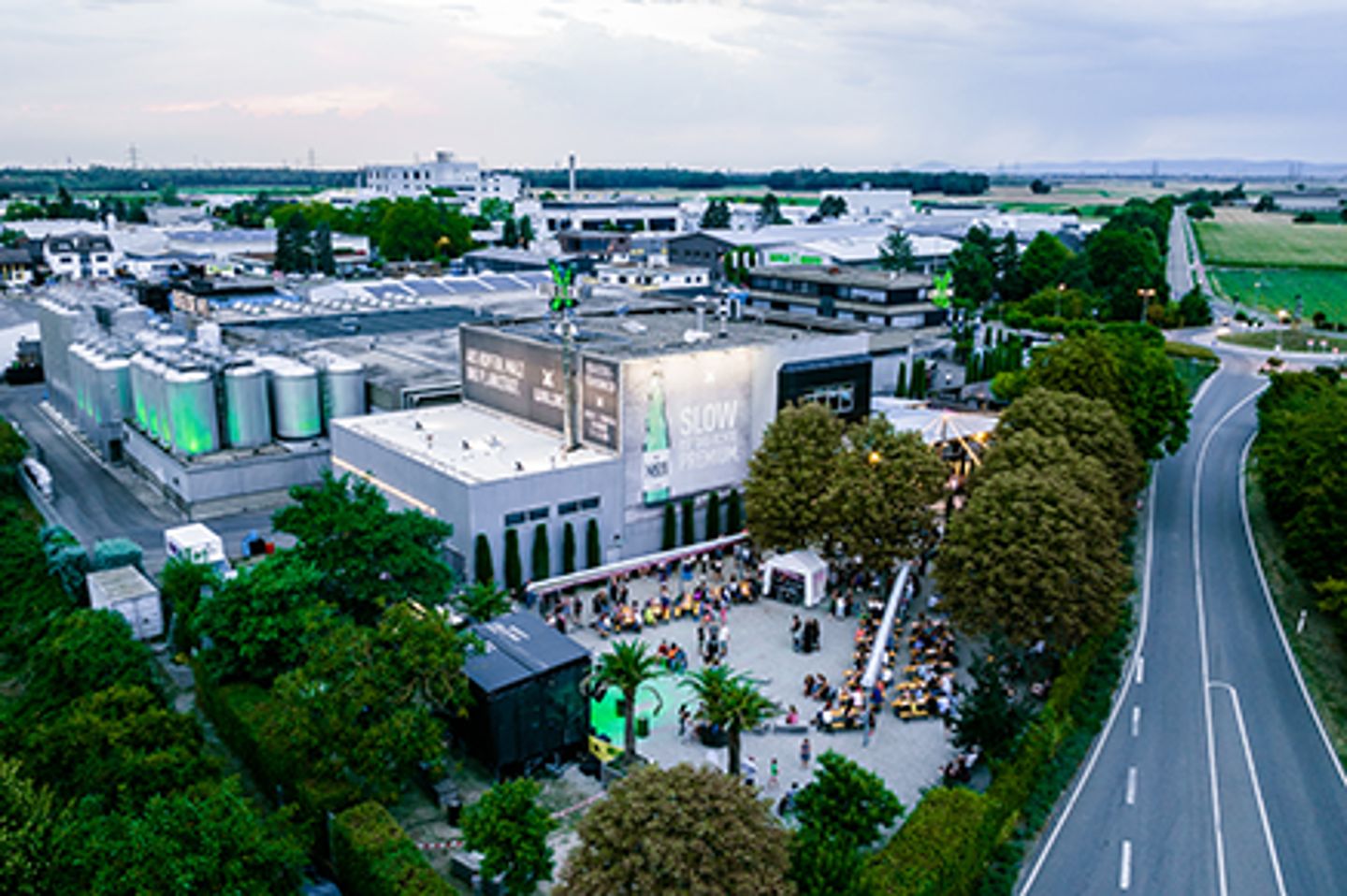 Aerial view of an industrial facility complex with outdoor gathering area and large green storage tanks at dusk
