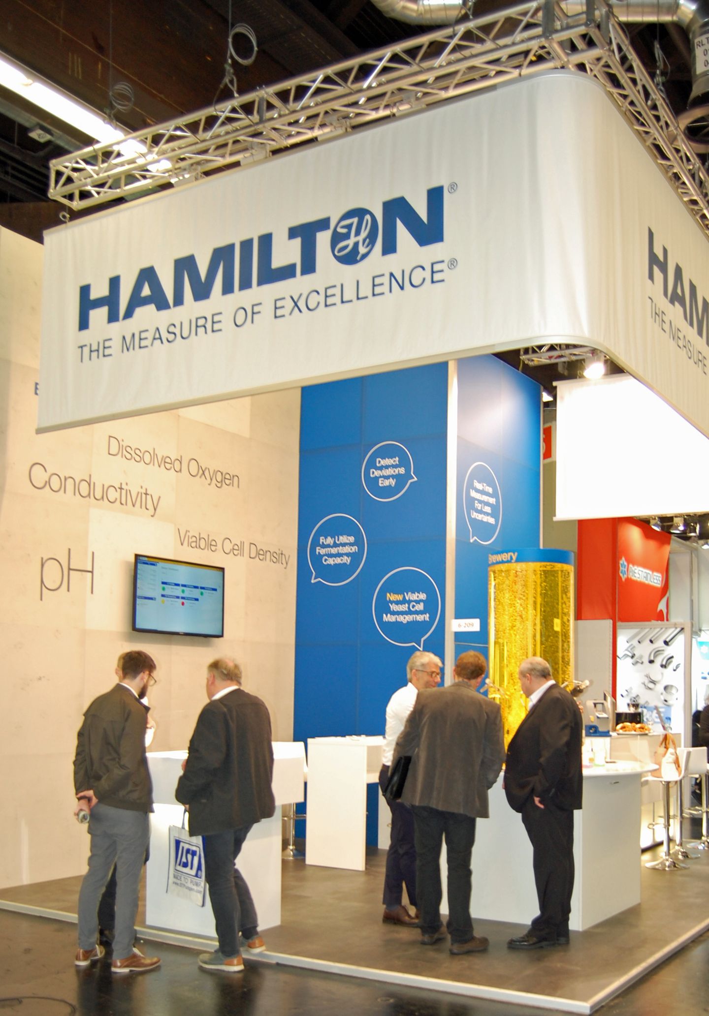 Hamilton "The Measure of Excellence" trade show booth displaying pH, conductivity, dissolved oxygen, and viable cell density process analytics, with attendees viewing a monitor and exhibition displays.