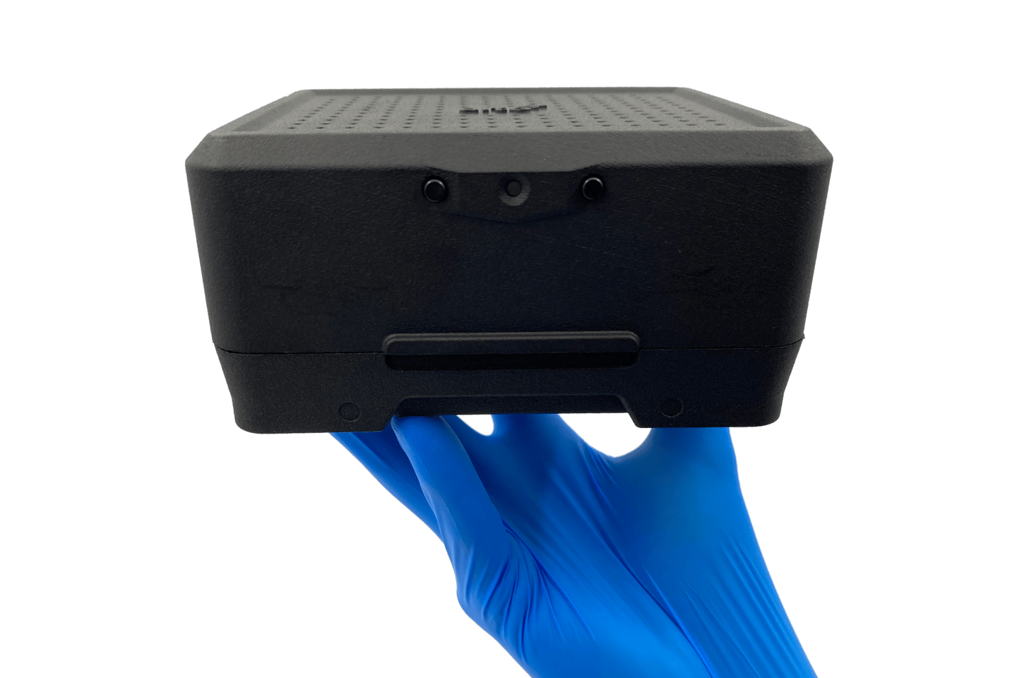 Gloved hand holding a compact black device with ventilation perforations on top and a slot on the front
