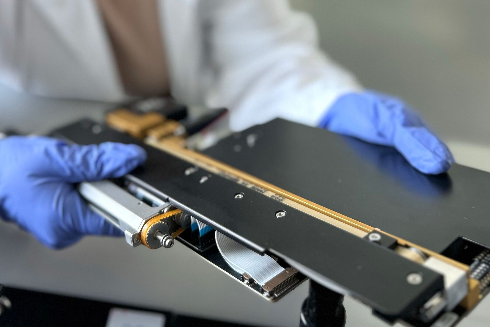 Gloved hands holding a precision linear module with gold rails, ribbon cables, and machined components
