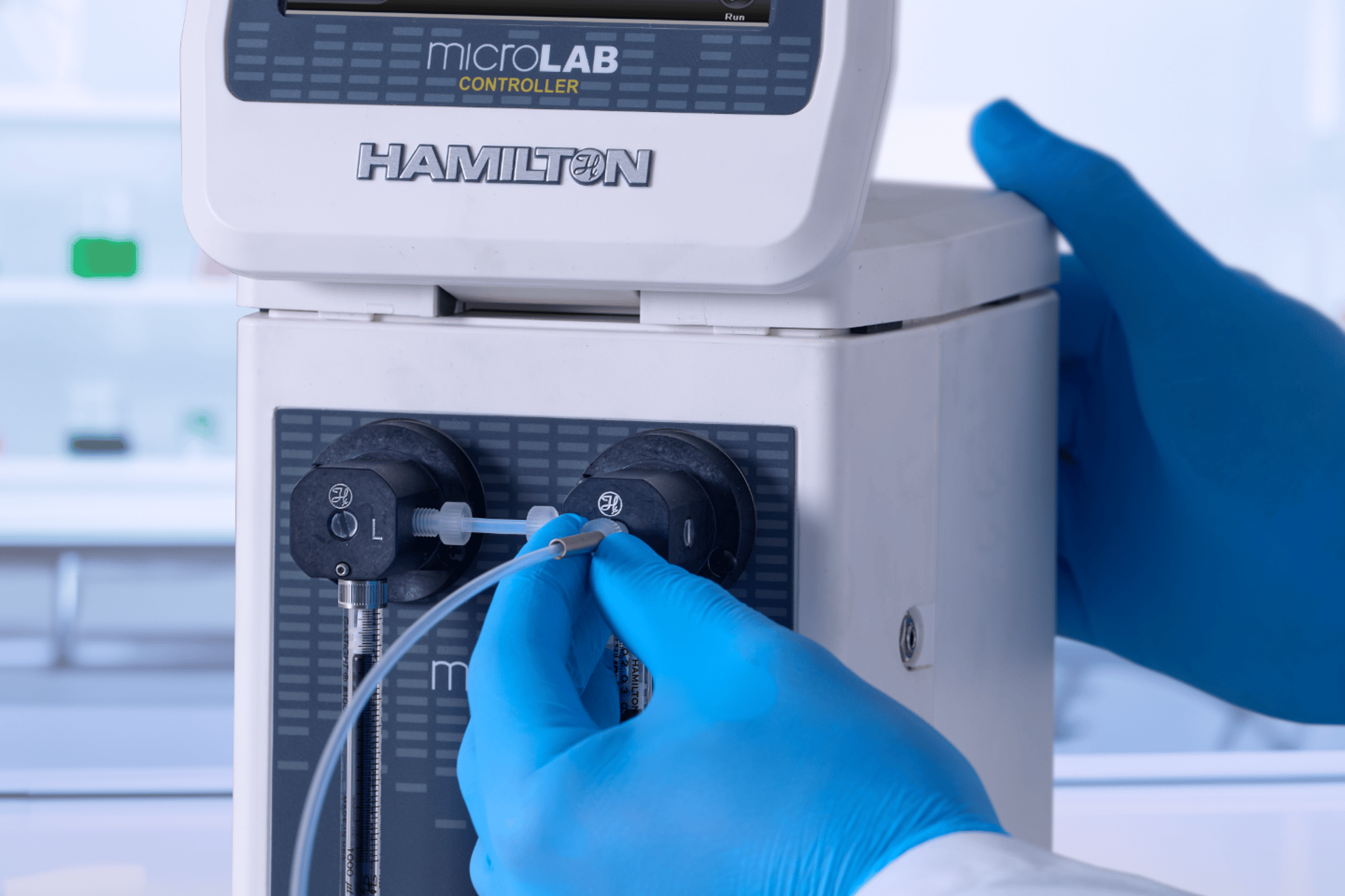 Gloved hands connecting tubing to a Hamilton MicroLab Controller syringe pump in a lab setting.