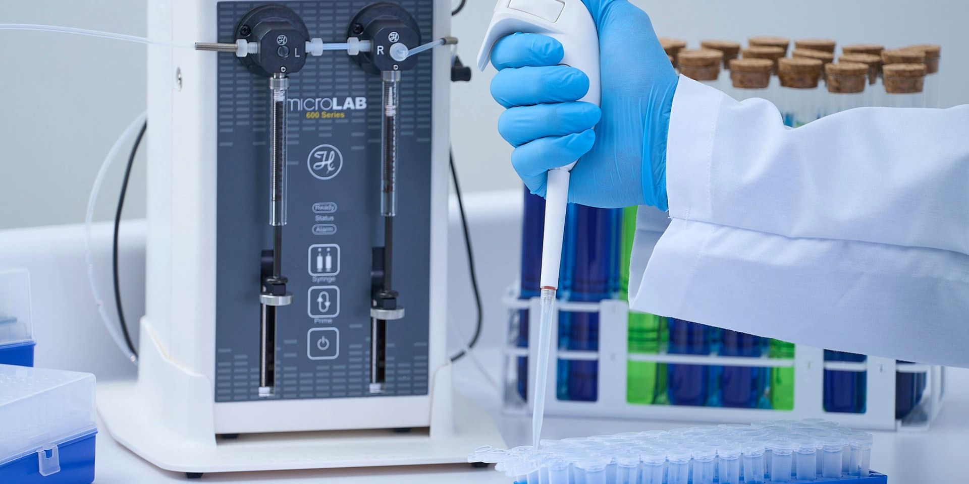 Gloved hand using a handheld pipette over a microplate beside a Hamilton MicroLab 600 Series dual-syringe dispenser