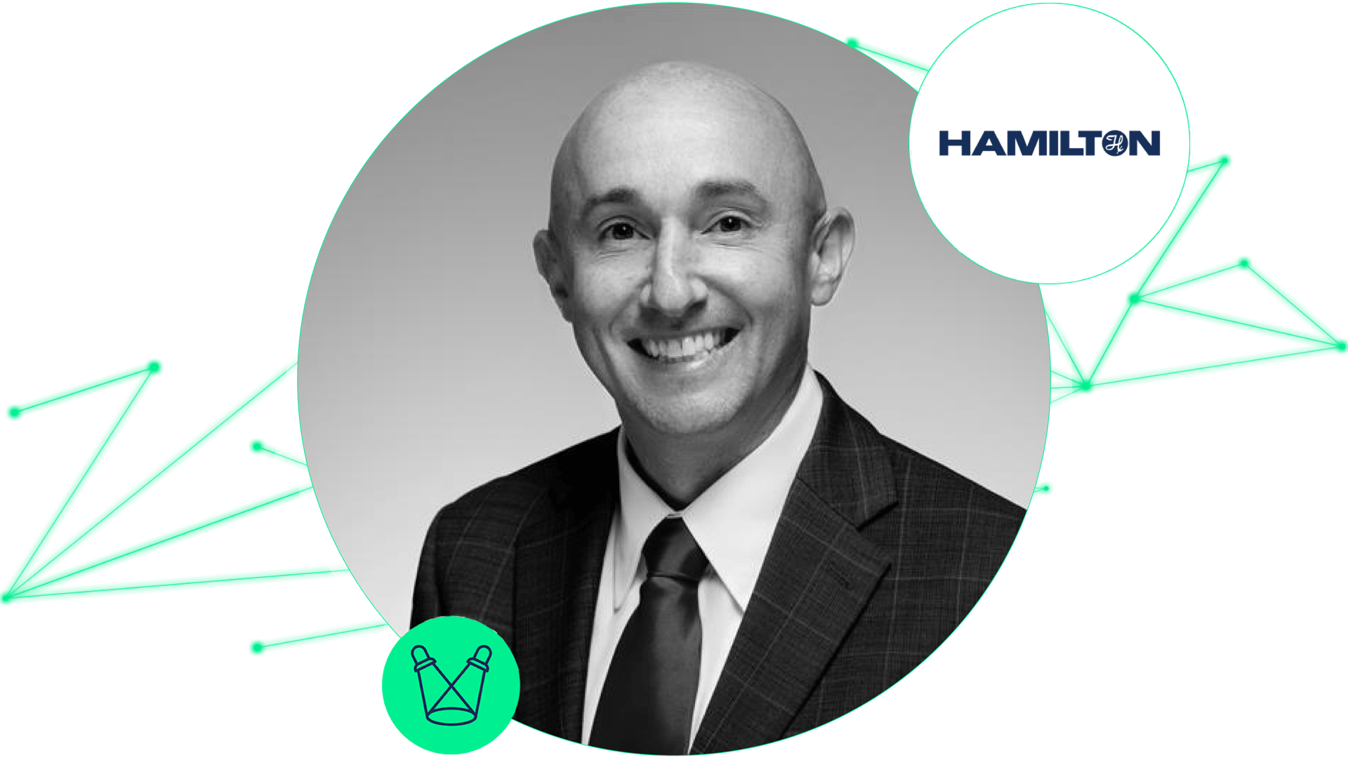 Smiling bald person in a dark plaid suit and tie, beside the Hamilton logo