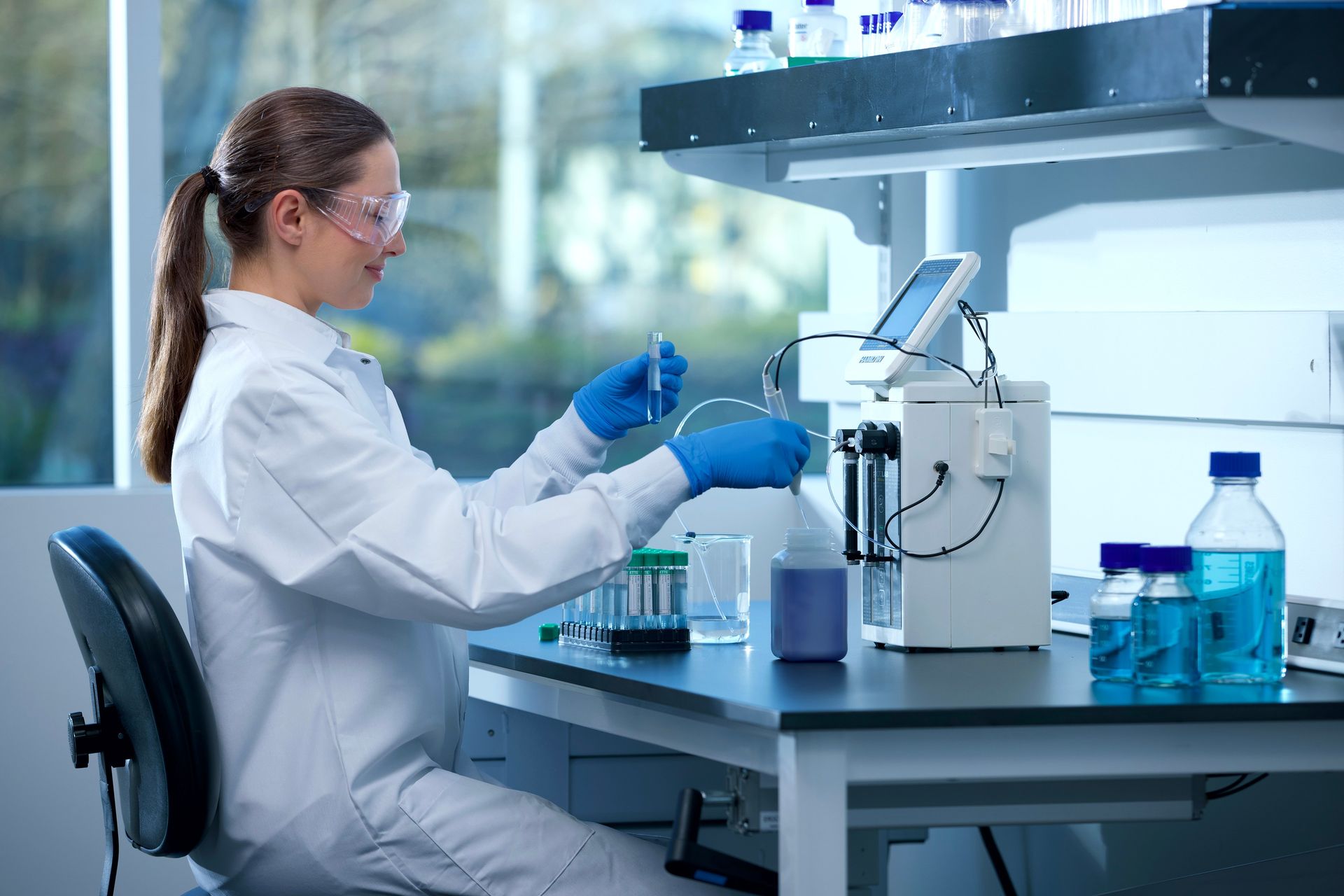 Lab professional in white coat and safety goggles pipetting samples near an analyzer with tubing, vials, and blue reagent bottles