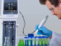 Researcher in gloves and safety glasses dispensing liquid into vials using a handheld dispenser, with a Hamilton microLAB 600 Series controller in the background.