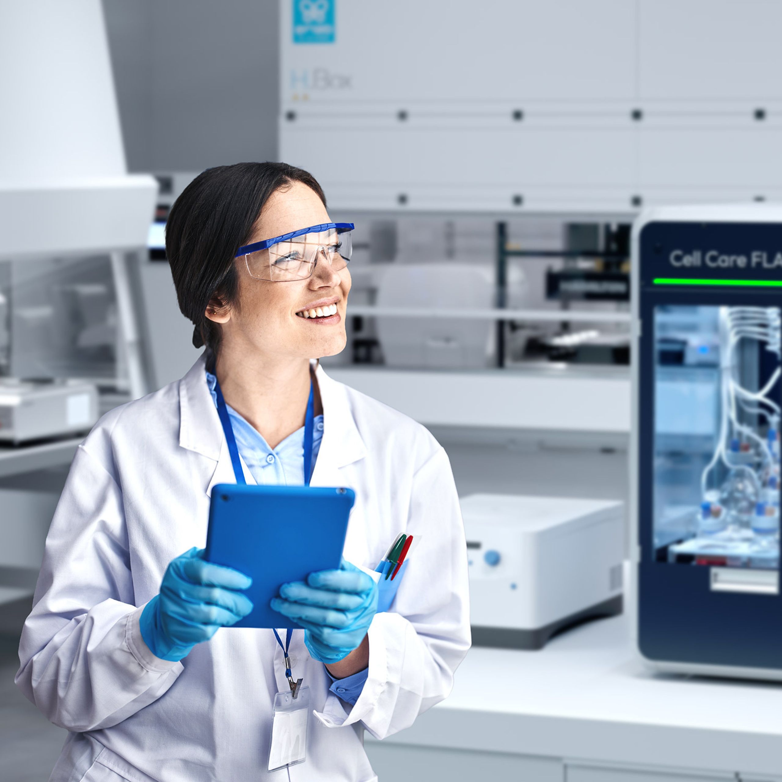 Smiling lab worker in white coat, safety glasses, and blue gloves holding a tablet in a modern laboratory