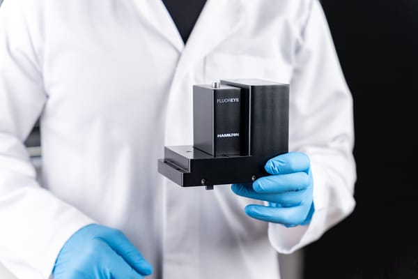 Lab-coated, gloved hands holding a black Hamilton FLUOREYE module against a dark background.