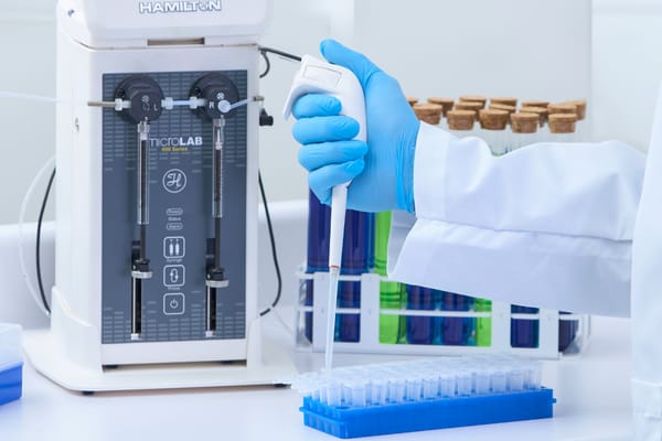 Gloved hand using an electronic pipette over a microplate, beside a Hamilton MicroLab 600 Series diluter