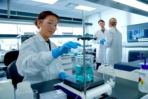 Three lab scientists in white coats and safety glasses working with beakers of blue liquid and lab instruments