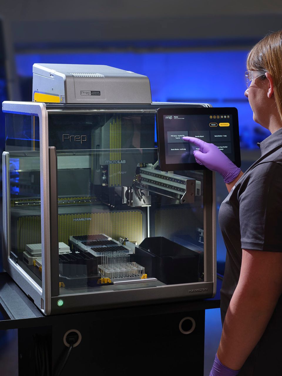 Lab technician in purple gloves operating a Hamilton MicroLab Prep automated liquid handler via touchscreen