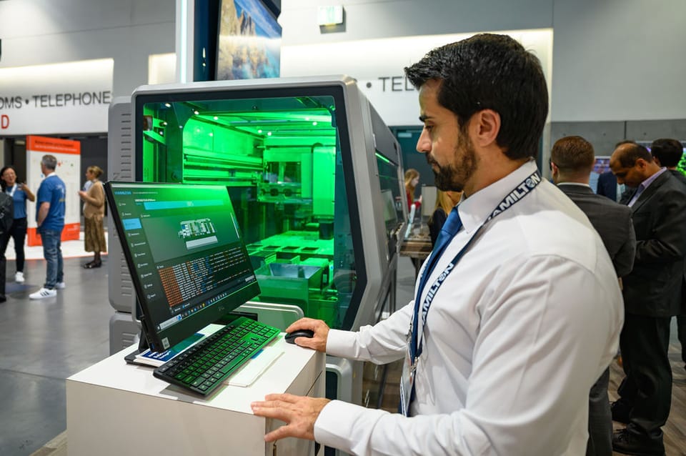 Hamilton representative operating a laptop connected to a green-lit automated liquid handling system at a trade show booth.