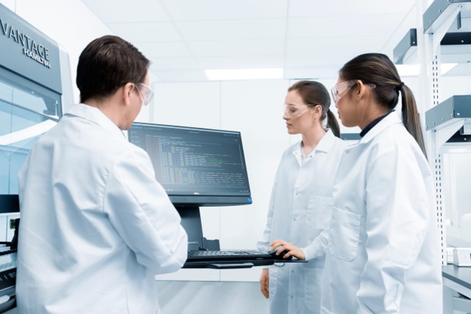 Three lab professionals in white coats and safety glasses review software on a monitor beside a Hamilton Vantage instrument