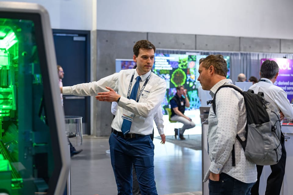 Hamilton representative gesturing toward a green-lit automated instrument while speaking with two attendees at a trade show booth.
