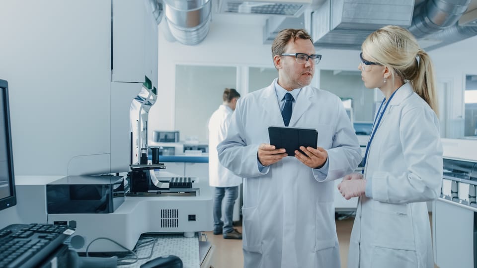 Two lab-coated researchers review a tablet beside automated laboratory instrumentation; a third colleague works in the background.
